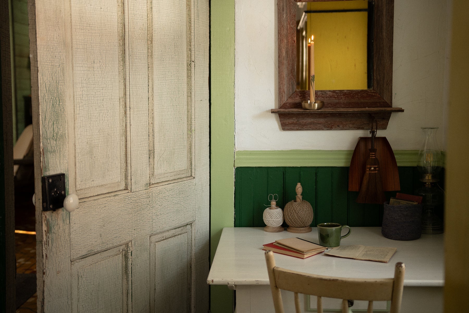 an antique desk, tucked away in the corner of a rustic homestead. the reflection in the mirror above the desk is of a yellow armoire in the background. a beeswax candle glows on the ledge above.