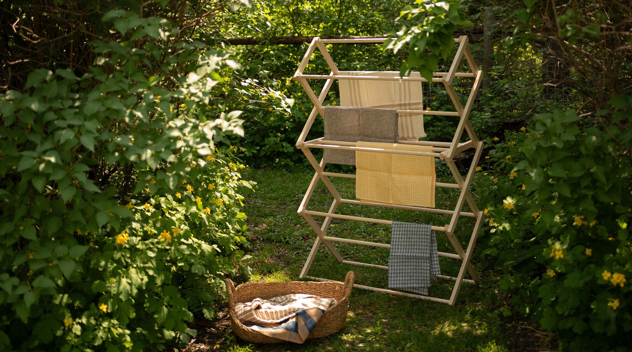 a wooden laundry drying rack with towels and a laundry basket dry under sundappled lilac trees