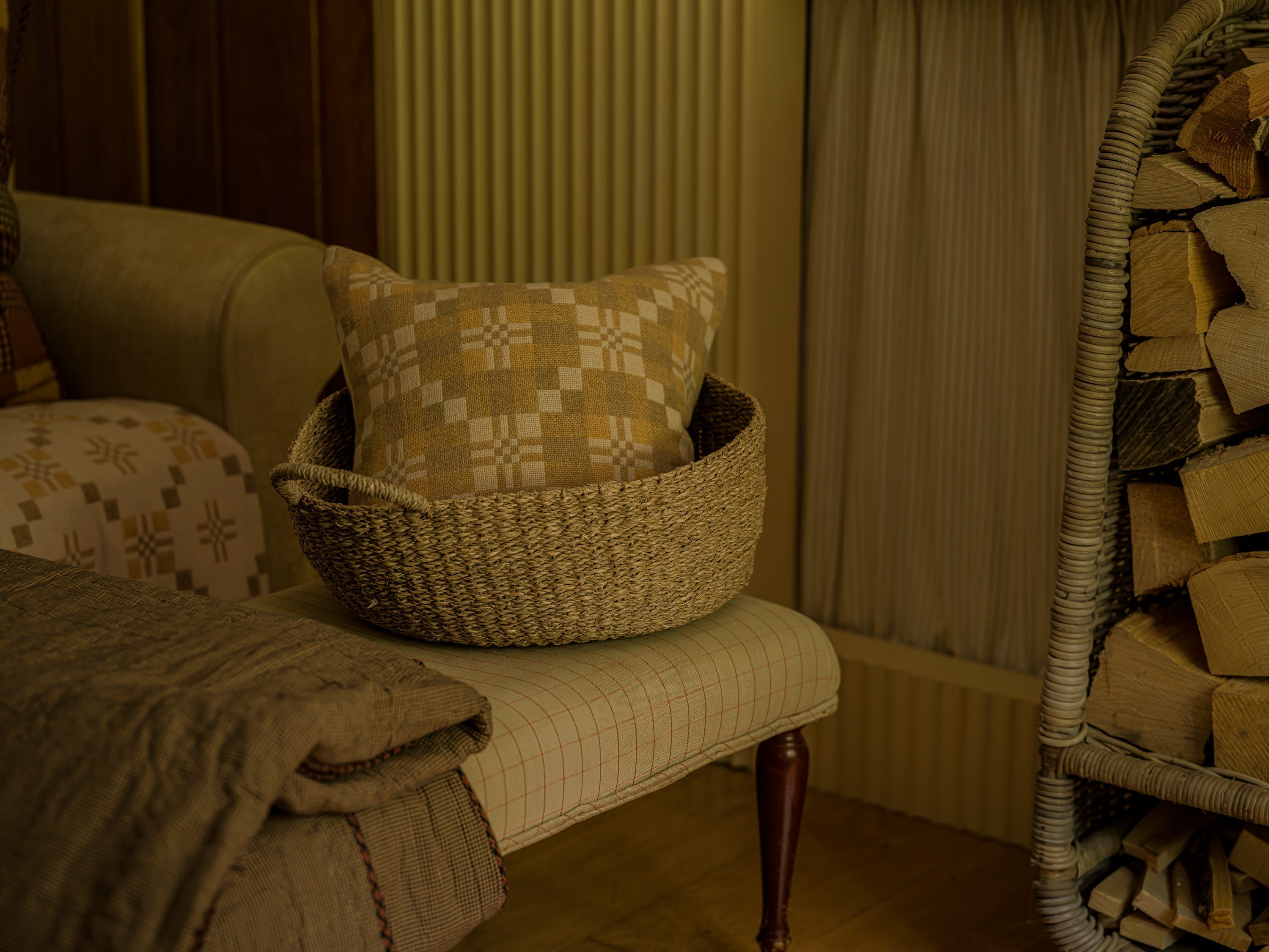 a plump wool throw cushion sitting in a seagrass basket atop an upholstered foot stool, near the fire, ready for movie night