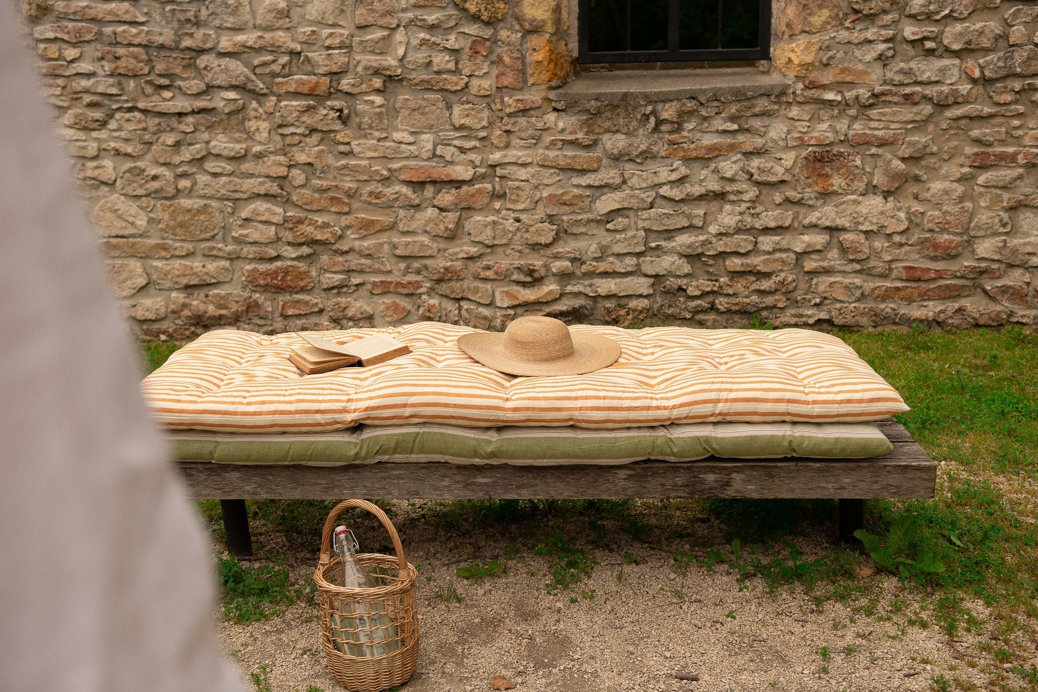 a wooden bench sits in front of a brick wall with cotton bedrolls, a sunhat, an open book, and a picnic basket