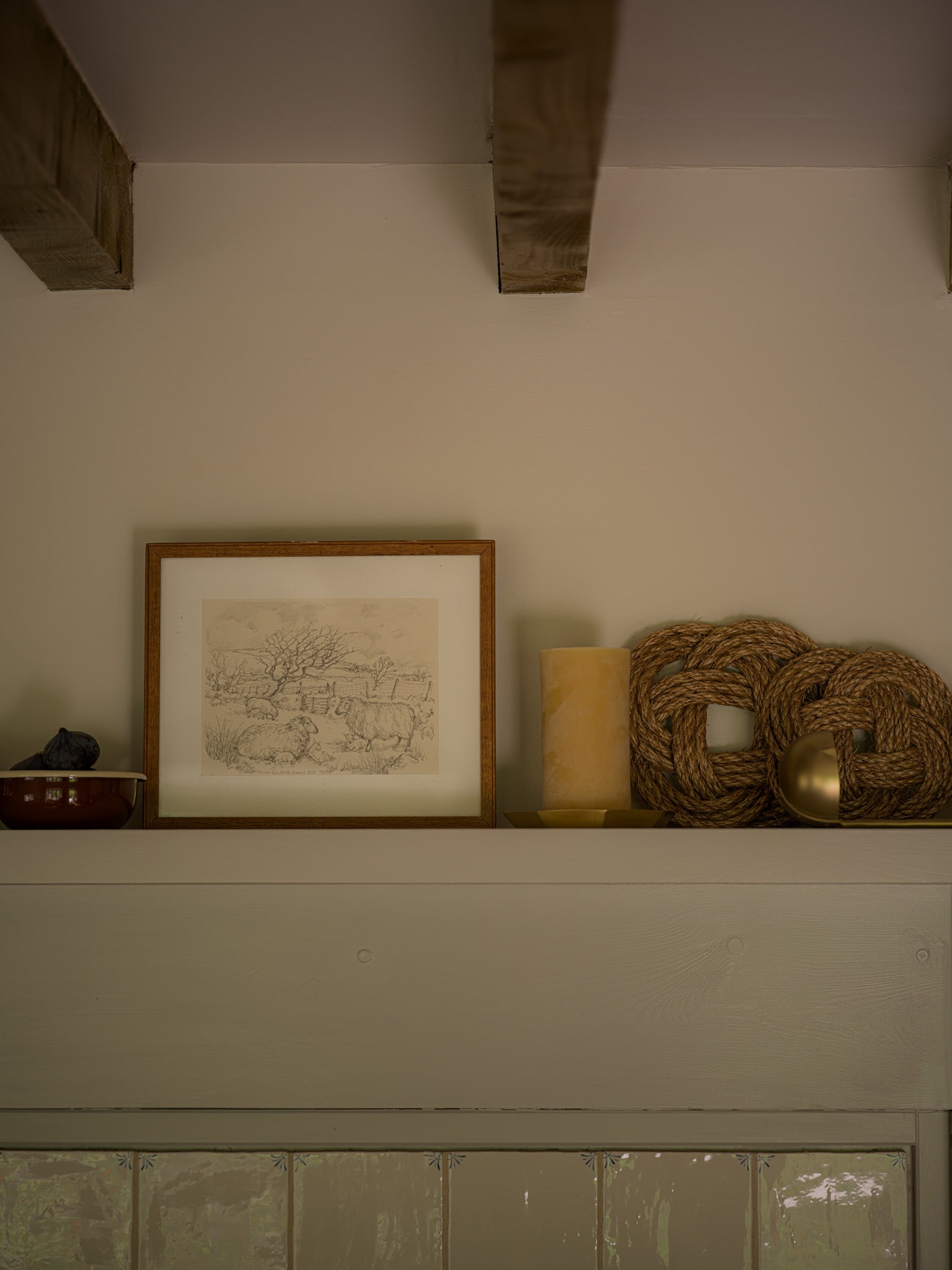 a framed sketch of two highland sheep sitting next to a beeswax candle and woven trivets on a ledge