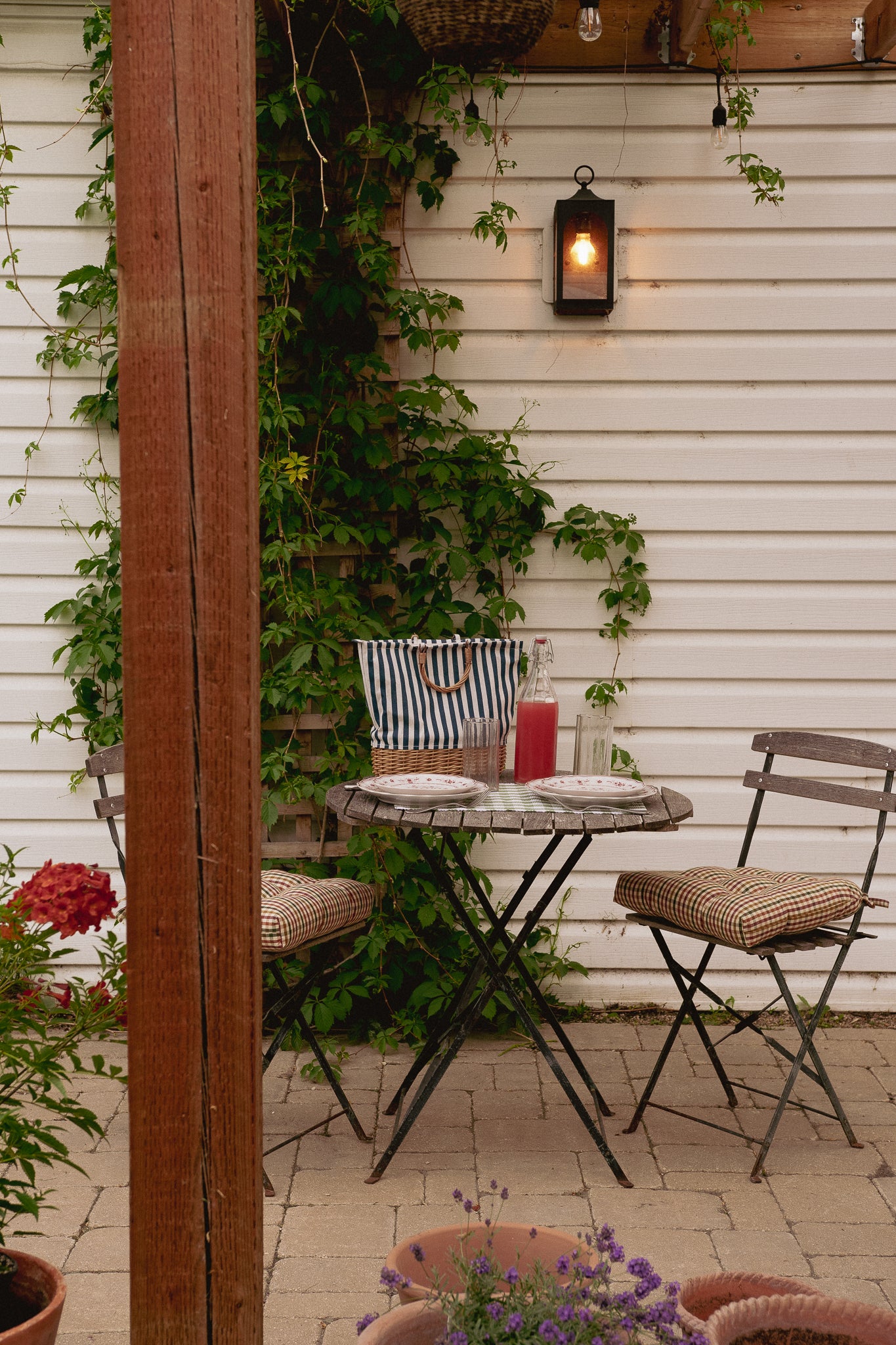 Outdoor patio dining setup with a small round table, two folding chairs with woven seats, a striped tote bag, and a hanging lantern against a white siding wall with green vines.