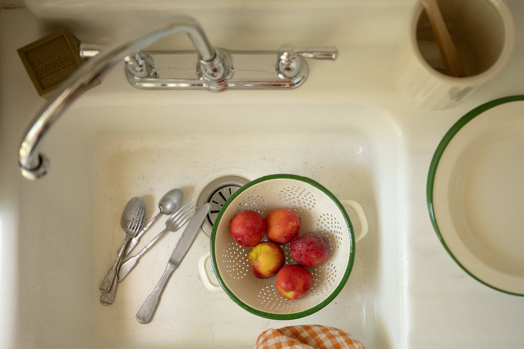 Peaches in a strainer inside a kitchen sink with utensils and a dishcloth.