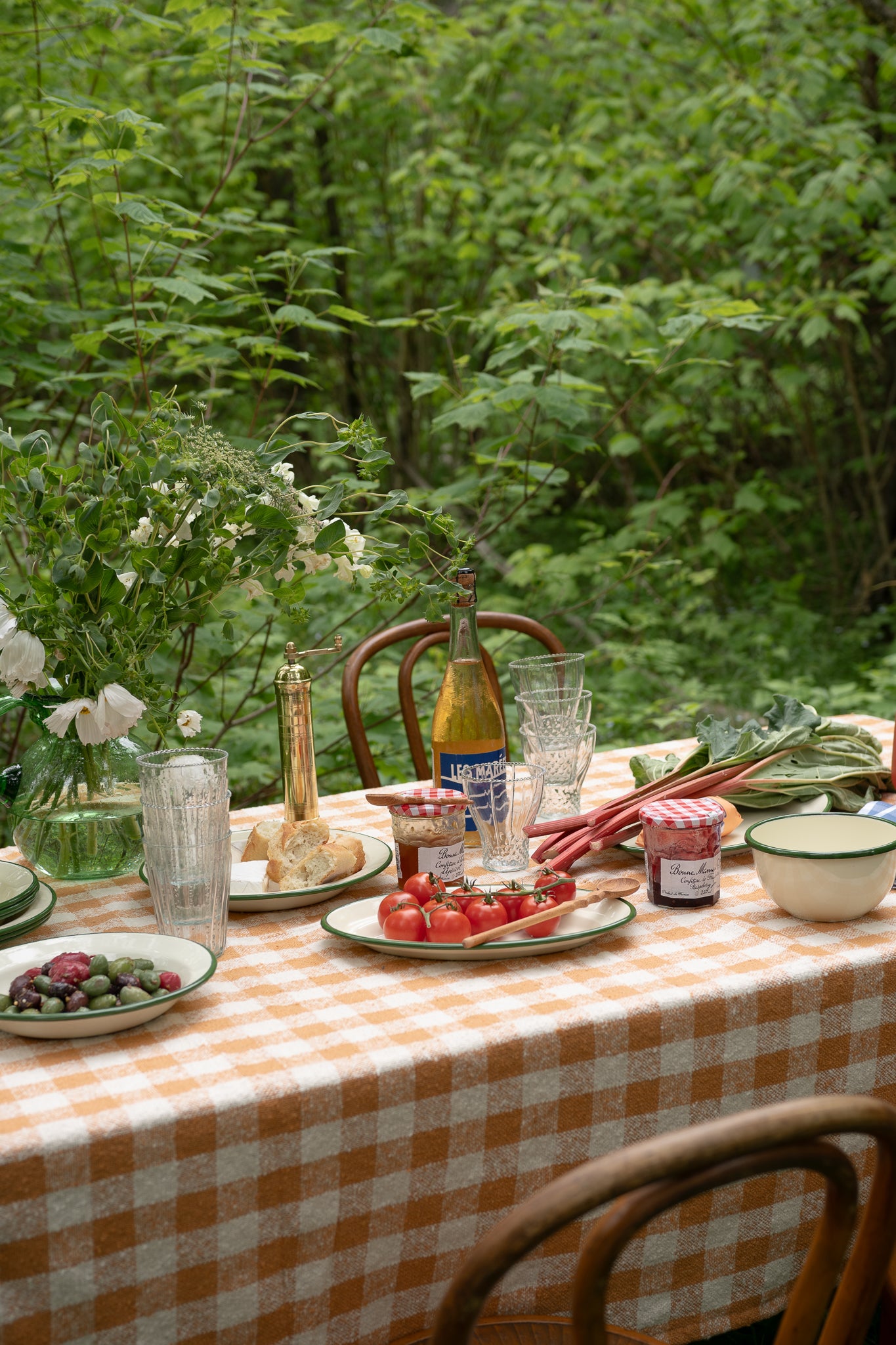 Outdoor dining table with checkered tablecloth, food, and drinks in a forest setting