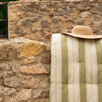 A striped quilted cushion or cushion cover in muted green and cream, draped over a rustic stone wall with a straw hat resting on top.