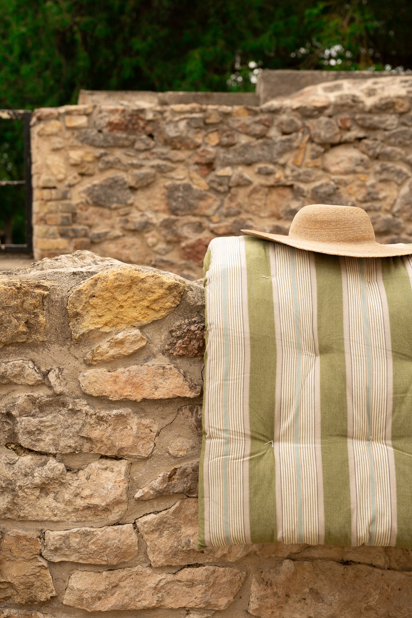 A striped quilted cushion or cushion cover in muted green and cream, draped over a rustic stone wall with a straw hat resting on top.