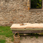 A rustic outdoor daybed with striped beige and orange cushions sits in front of a stone wall, with a small woven basket on the sandy ground nearby.