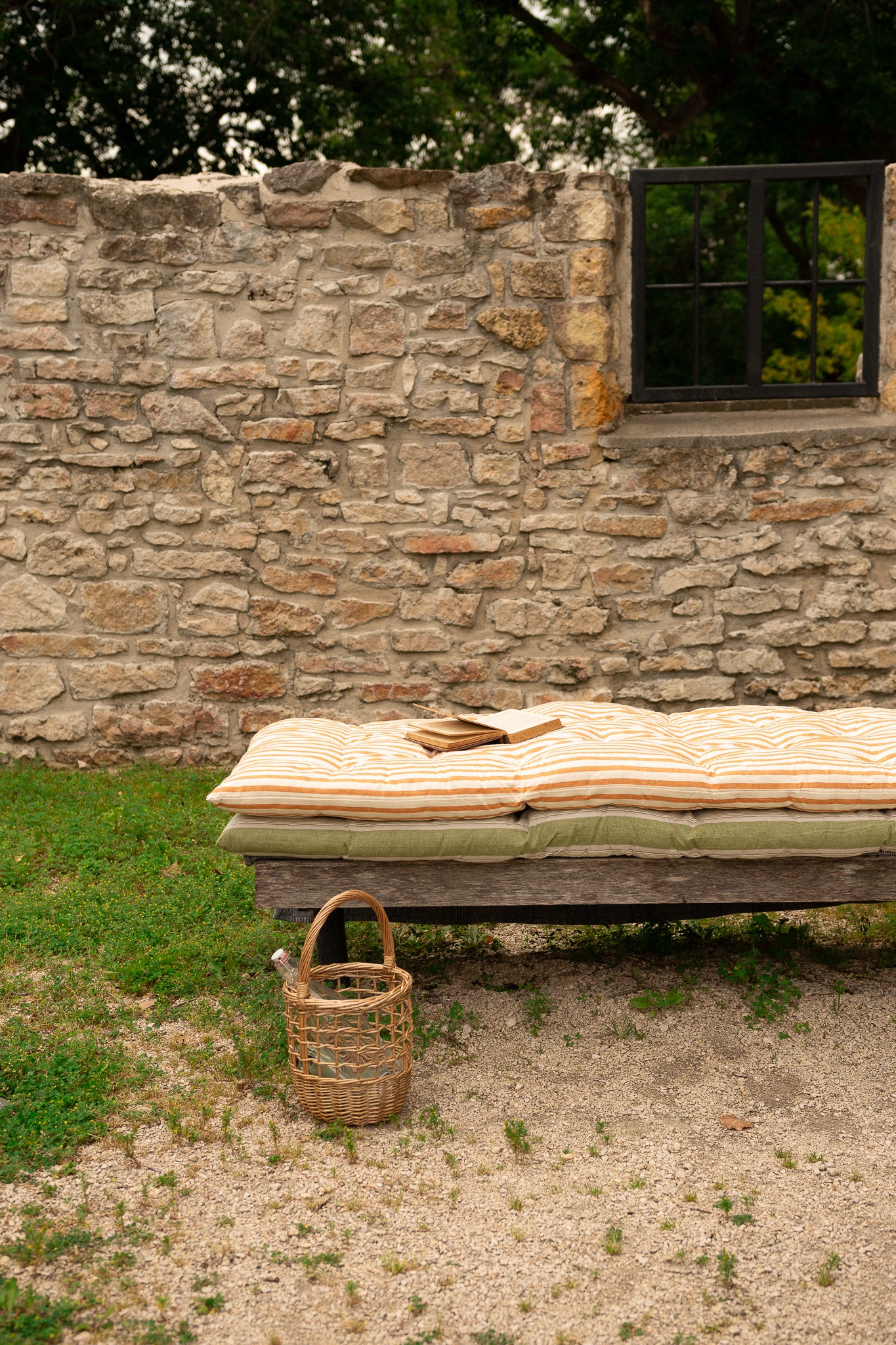 A rustic outdoor daybed with striped beige and orange cushions sits in front of a stone wall, with a small woven basket on the sandy ground nearby.
