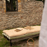 A rustic outdoor daybed with striped cream and tan cushions, set against a stone wall with a wicker basket and straw hat nearby.