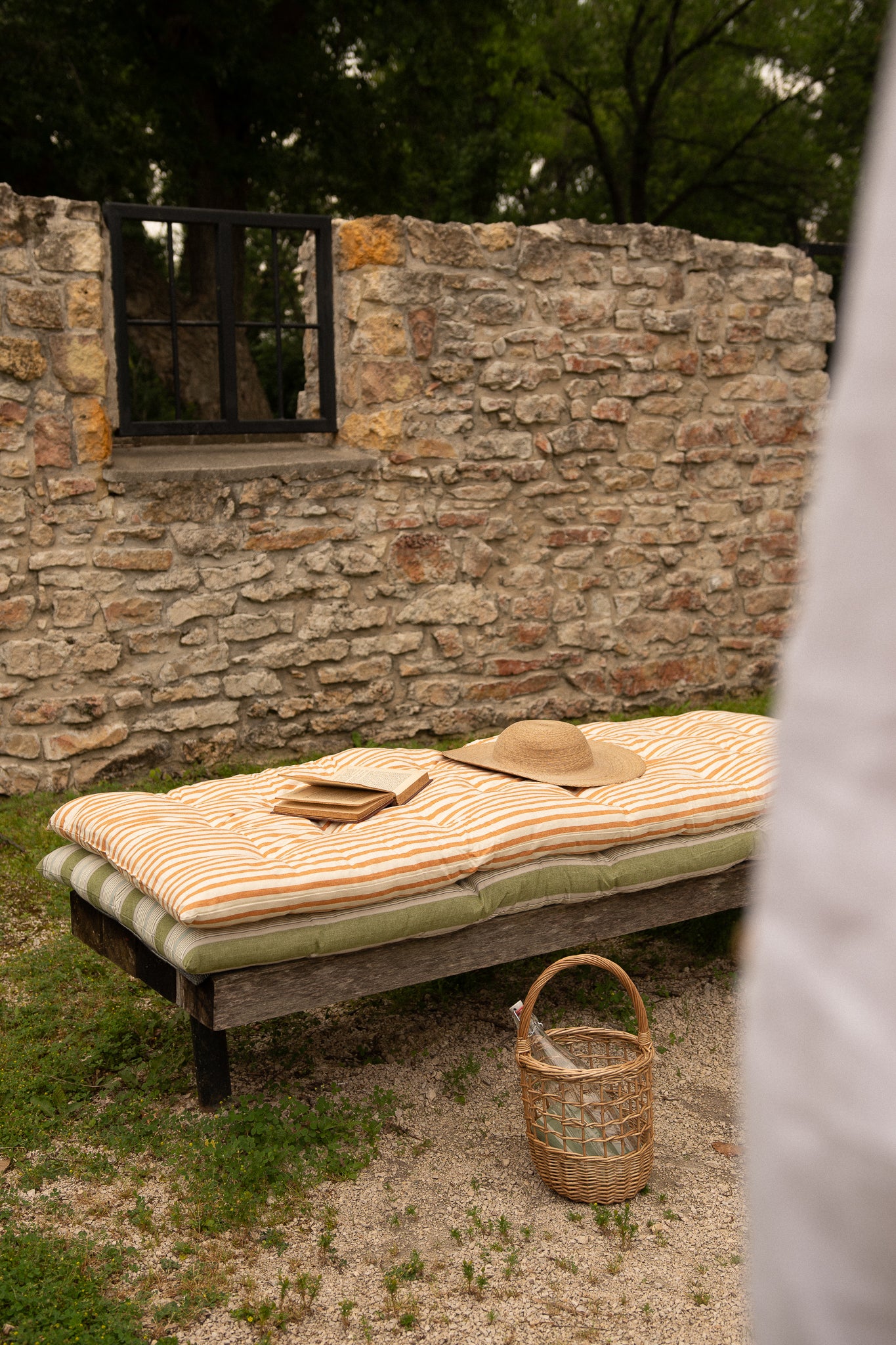 A rustic outdoor daybed with striped cream and tan cushions, set against a stone wall with a wicker basket and straw hat nearby.