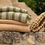 Folded striped and checkered cushions stacked on a rustic stone wall, with an outdoor stone path and greenery blurred in the background.