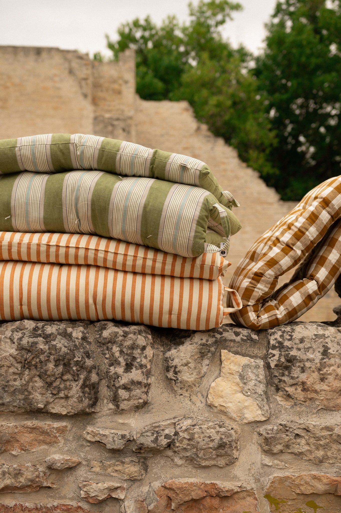 Folded striped and checkered cushions stacked on a rustic stone wall, with an outdoor stone path and greenery blurred in the background.