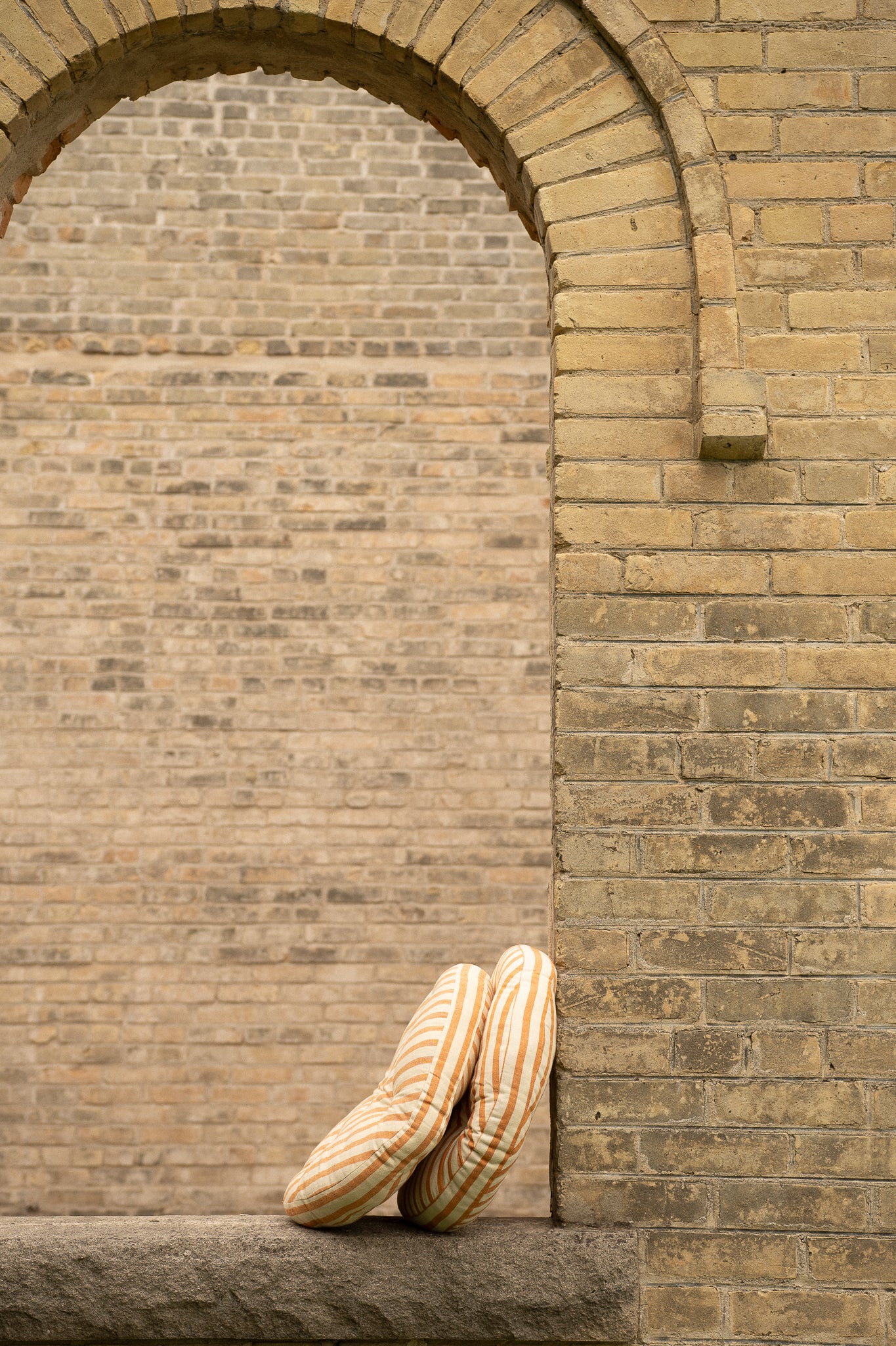 A pair of beige and white striped knit slip-on slippers resting on a stone ledge against a tan brick wall with an arched opening.