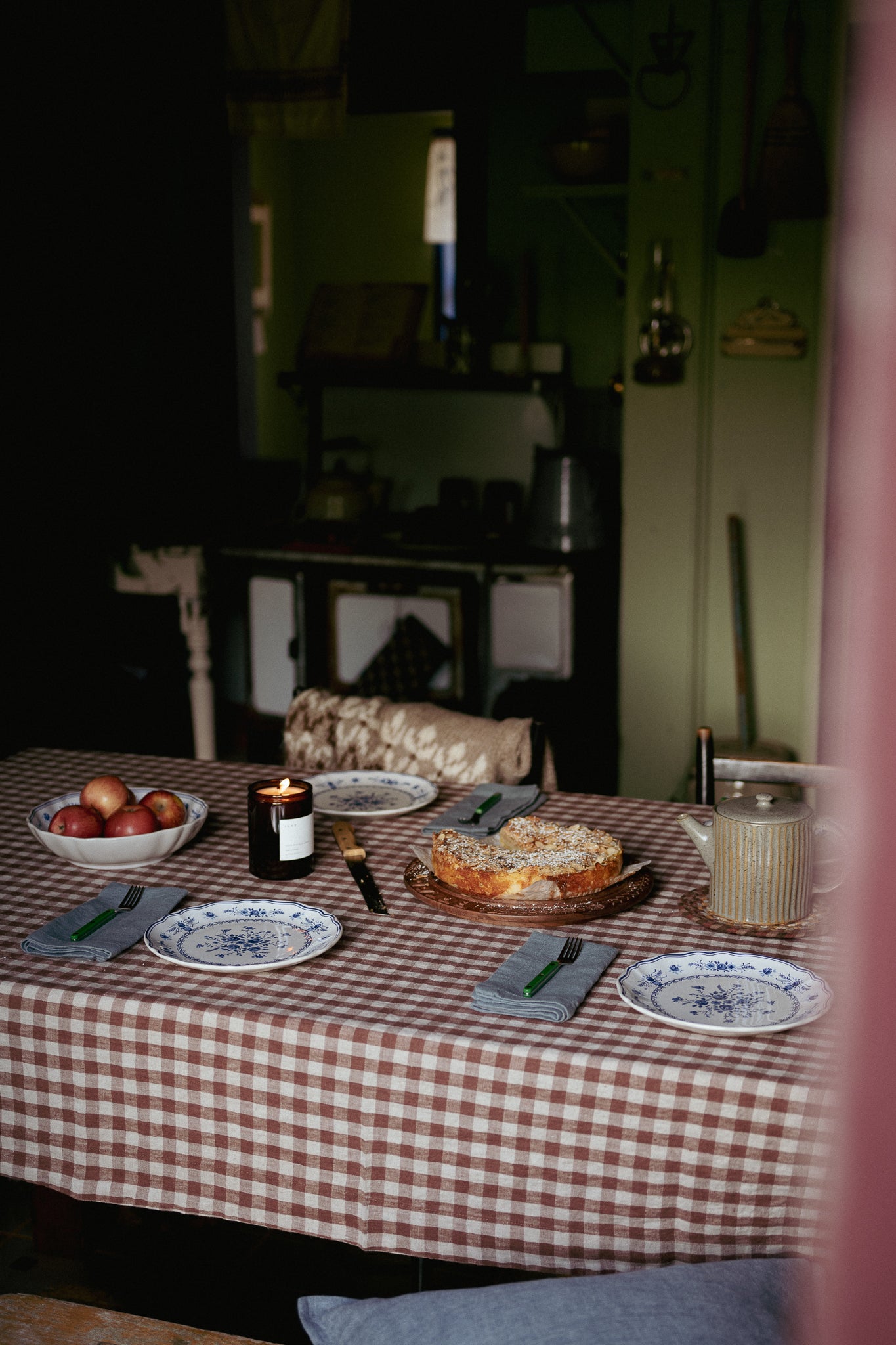 A rustic dining table set in a dimly lit kitchen or dining room, featuring a red and white checkered tablecloth, blue and white patterned plates, a lit candle, a bowl of red apples, and a sliced cake on a wooden board.