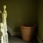 A natural woven wicker storage basket with two handles sits on a dark gray staircase landing against olive green walls, next to a white wooden banister.