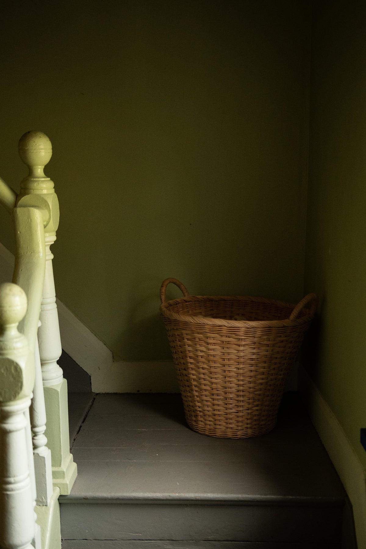 A natural woven wicker storage basket with two handles sits on a dark gray staircase landing against olive green walls, next to a white wooden banister.