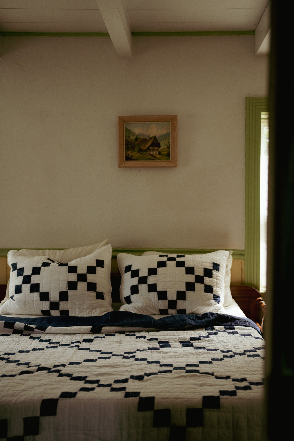 A bed dressed in a black and white geometric patterned quilt and matching pillows, set in a rustic bedroom with a small landscape painting on the wall above.
