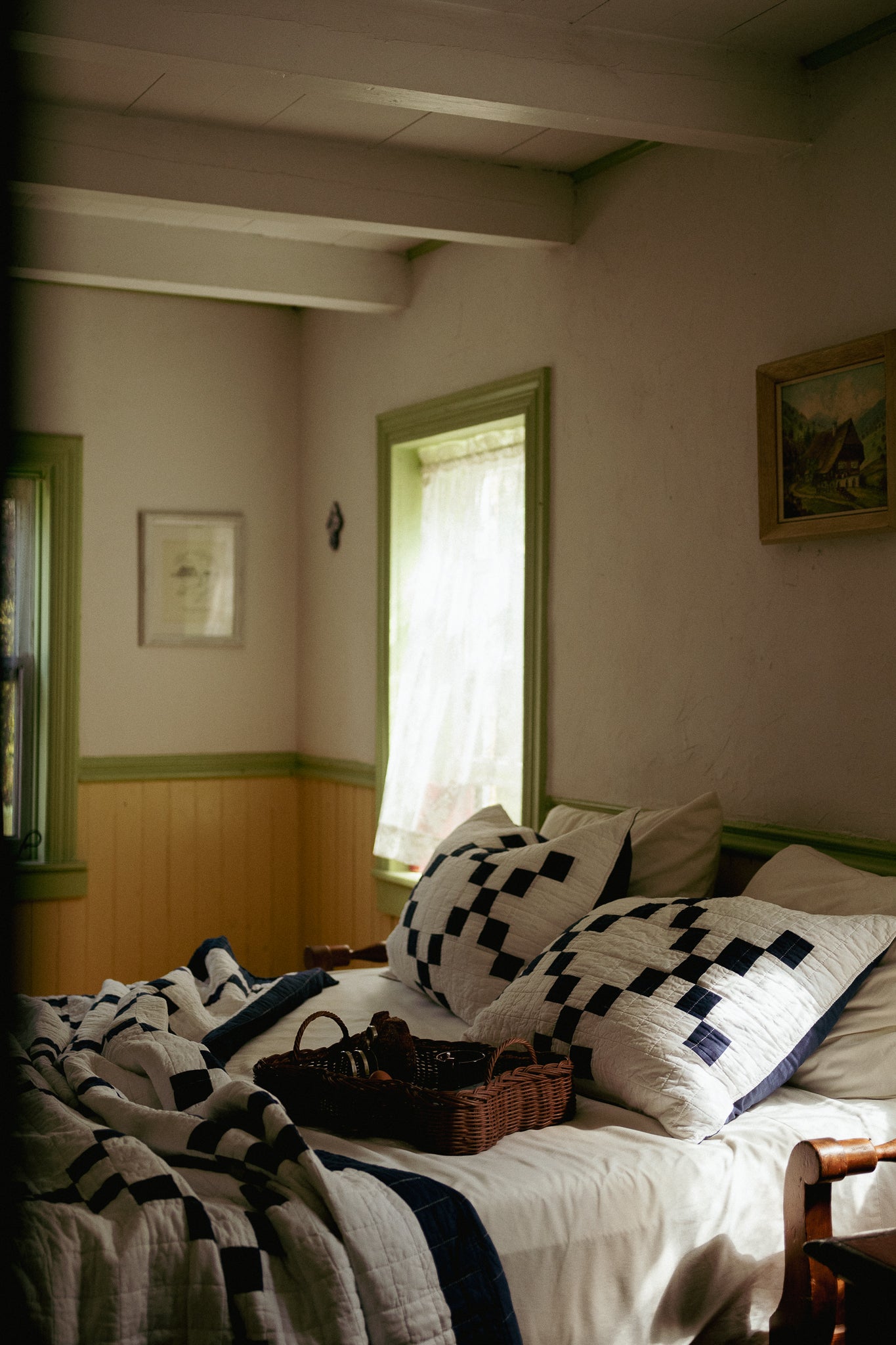 A rustic bedroom scene with a bed dressed in white bedding and black-and-white checkered pillows and quilt, set in a dimly lit room with green-trimmed walls and windows.