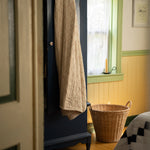 A dark navy blue wooden armoire or cabinet in a cozy room, with a beige quilted throw blanket draped over its door. A wicker basket sits on the floor beside a window with green trim, and part of a patterned quilt is visible in the foreground.