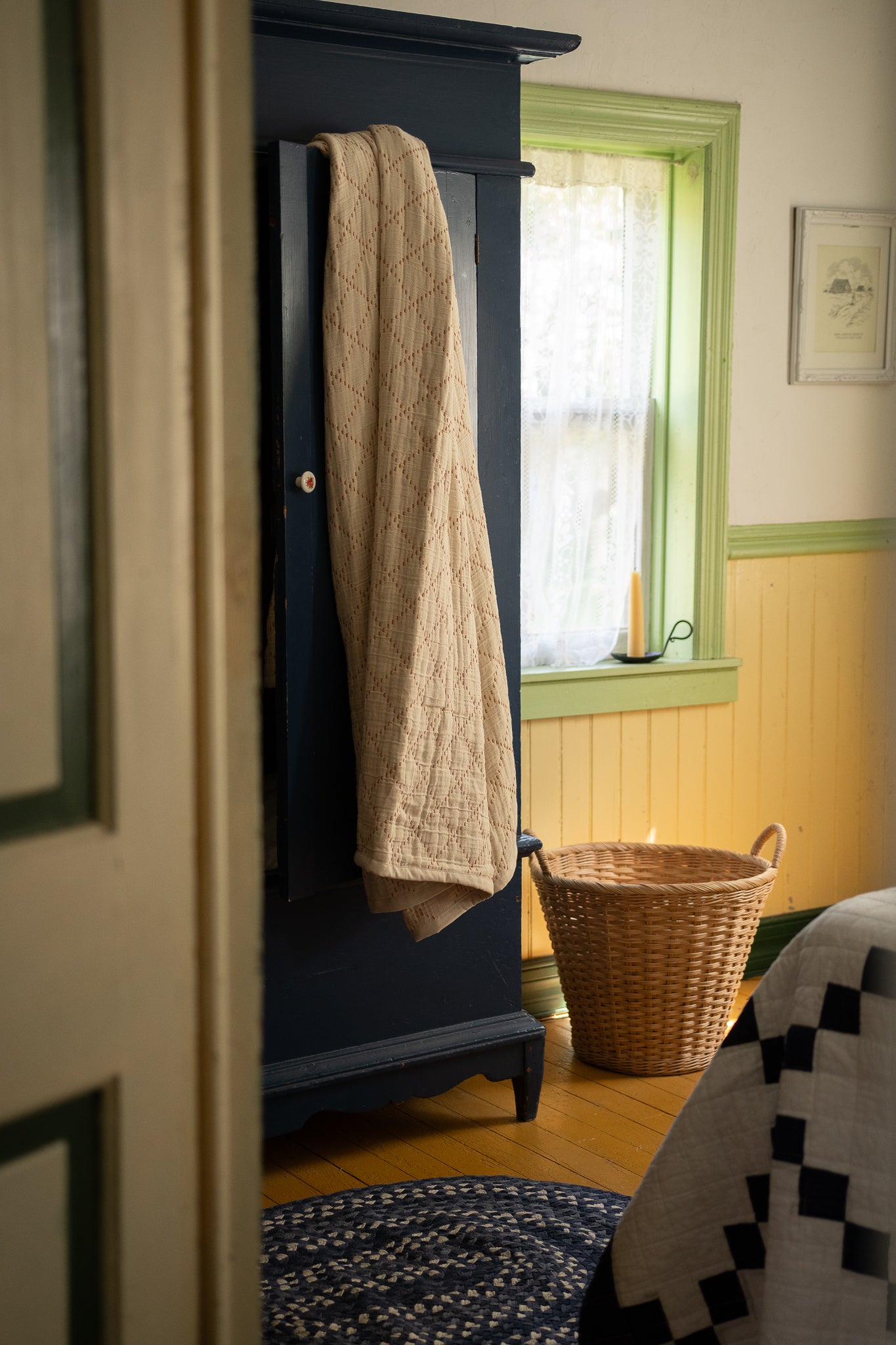 A dark navy blue wooden armoire or cabinet in a cozy room, with a beige quilted throw blanket draped over its door. A wicker basket sits on the floor beside a window with green trim, and part of a patterned quilt is visible in the foreground.