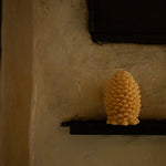 A textured, honeycomb-like yellow candle shaped like a pine cone, displayed on a dark shelf against a warm, textured beige plaster wall.