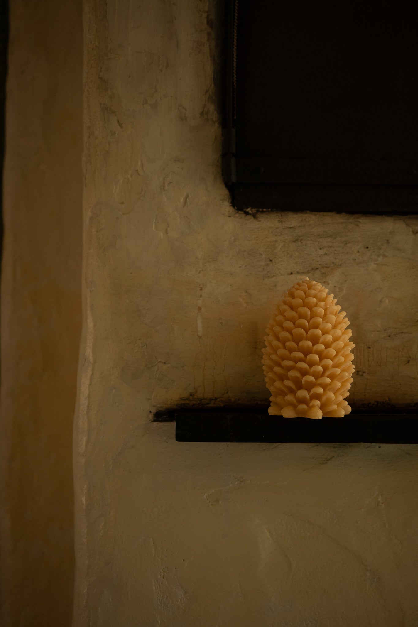 A textured, honeycomb-like yellow candle shaped like a pine cone, displayed on a dark shelf against a warm, textured beige plaster wall.
