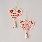 Two heart-shaped embroidered fabric ornaments in cream and red hanging on a light gray background. One ornament has a floral pattern and the other has a heart-and-heart medallion design, both with small tassels and gold hanging strings.