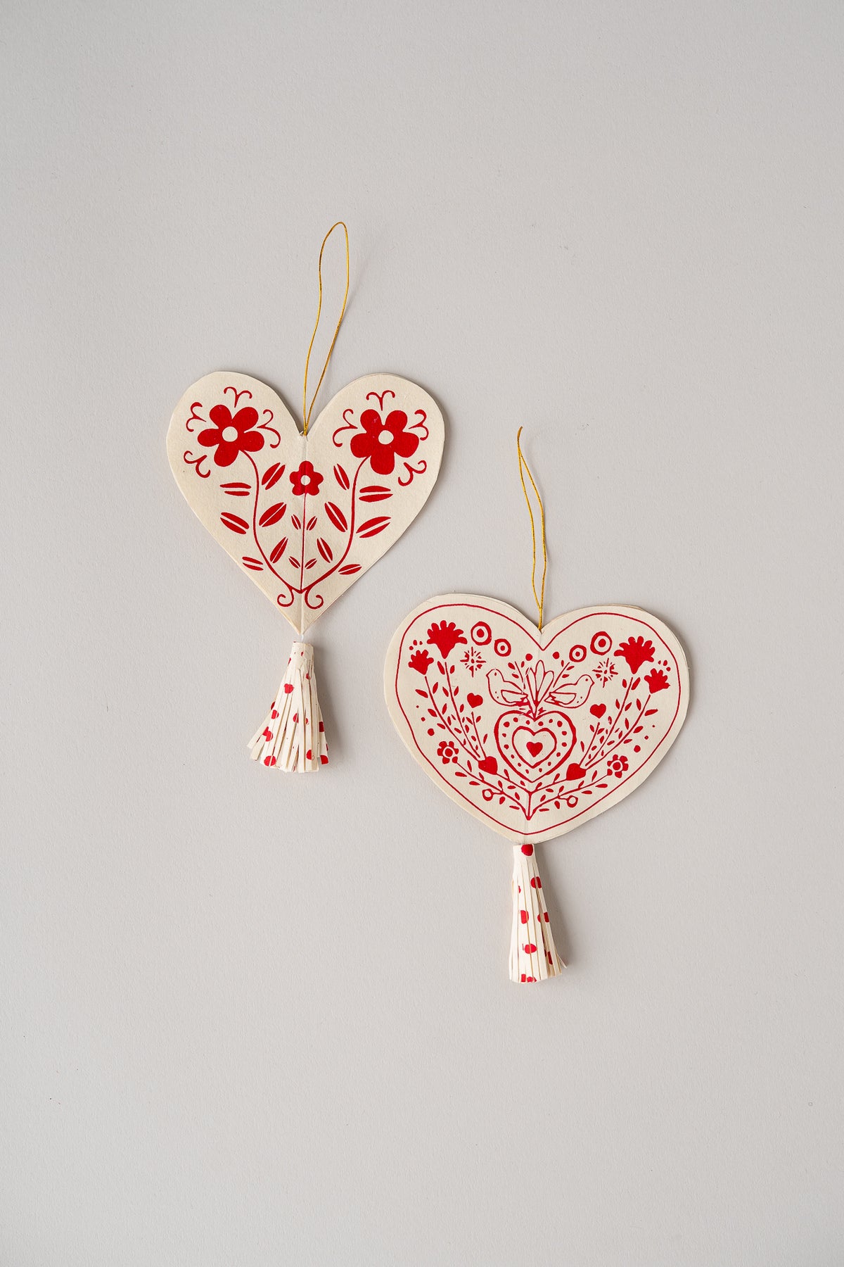 Two heart-shaped embroidered fabric ornaments in cream and red hanging on a light gray background. One ornament has a floral pattern and the other has a heart-and-heart medallion design, both with small tassels and gold hanging strings.