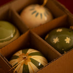 A close-up of four hand-painted ceramic Christmas ornaments arranged in compartments of a cardboard box. The ornaments feature green, cream, and gold patterns including stripes, flowers, and dots.