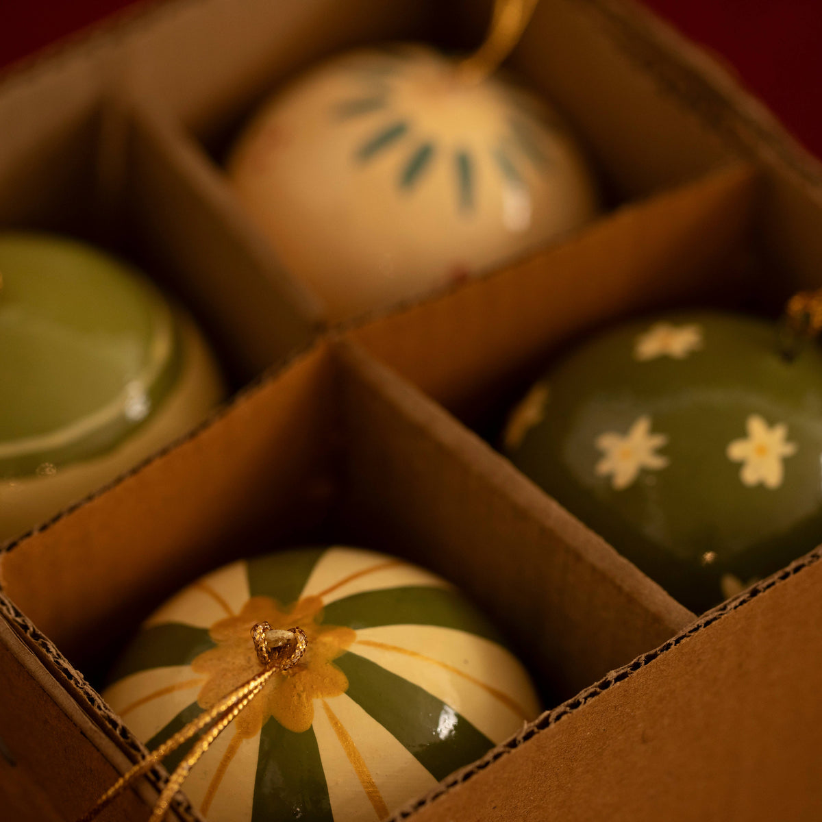 A close-up of four hand-painted ceramic Christmas ornaments arranged in compartments of a cardboard box. The ornaments feature green, cream, and gold patterns including stripes, flowers, and dots.
