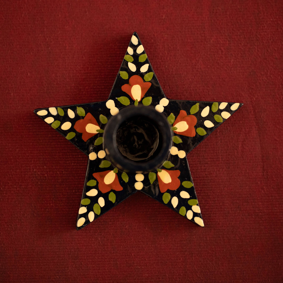 A five-pointed star-shaped black candle holder with hand-painted floral and leaf patterns in red, green, and cream, set against a dark red textured background.
