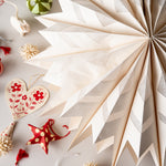 A large cream-colored paper fan decoration with a geometric zigzag pattern, displayed on a light gray surface alongside other festive ornaments and decorative items.