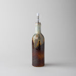 A decorative glass bottle with a spout, featuring a gradient of amber-brown at the bottom, dark brown speckling in the middle, and a light blue-green top section, set against a solid light gray background.