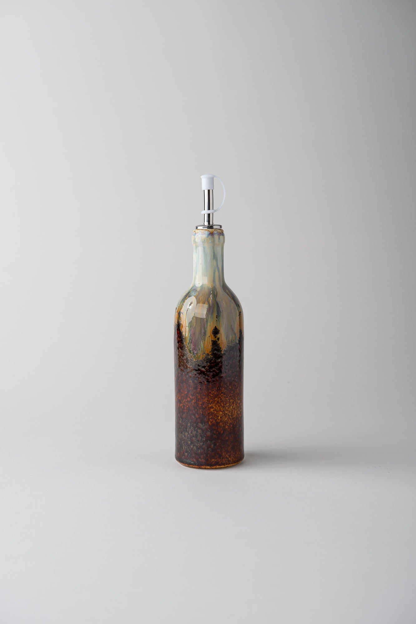 A decorative glass bottle with a spout, featuring a gradient of amber-brown at the bottom, dark brown speckling in the middle, and a light blue-green top section, set against a solid light gray background.