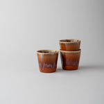 Three small glazed ceramic cups with a brown and amber drip design, arranged on a light gray background.