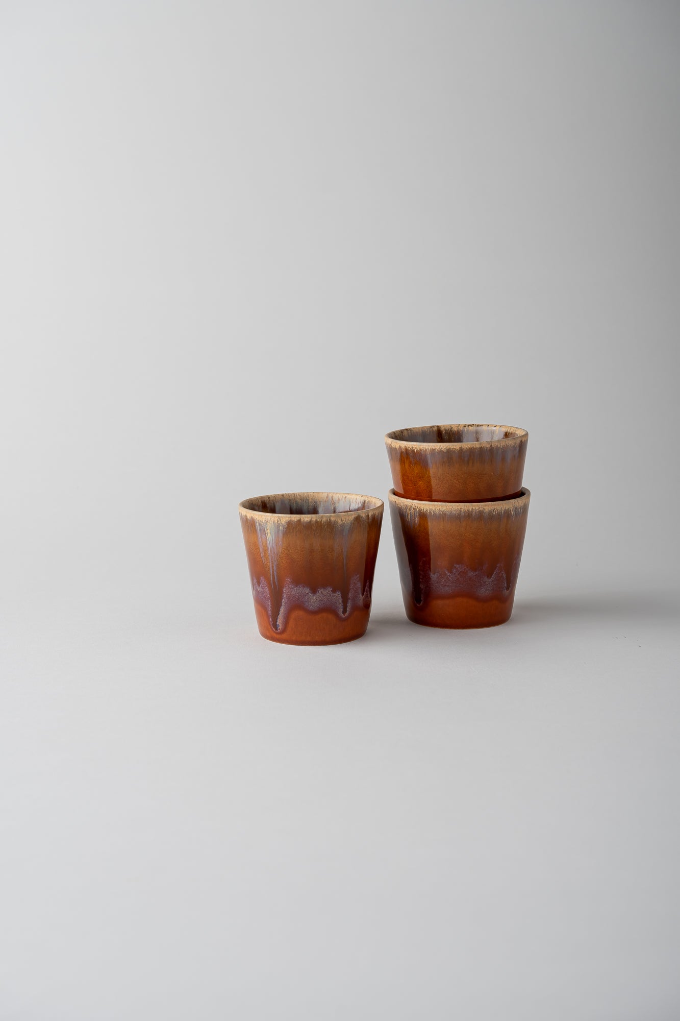 Three small glazed ceramic cups with a brown and amber drip design, arranged on a light gray background.