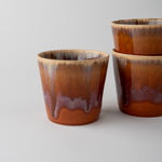 Three small ceramic cups with a glossy brown and amber glaze, featuring a dripping effect near the base, arranged on a light gray background.