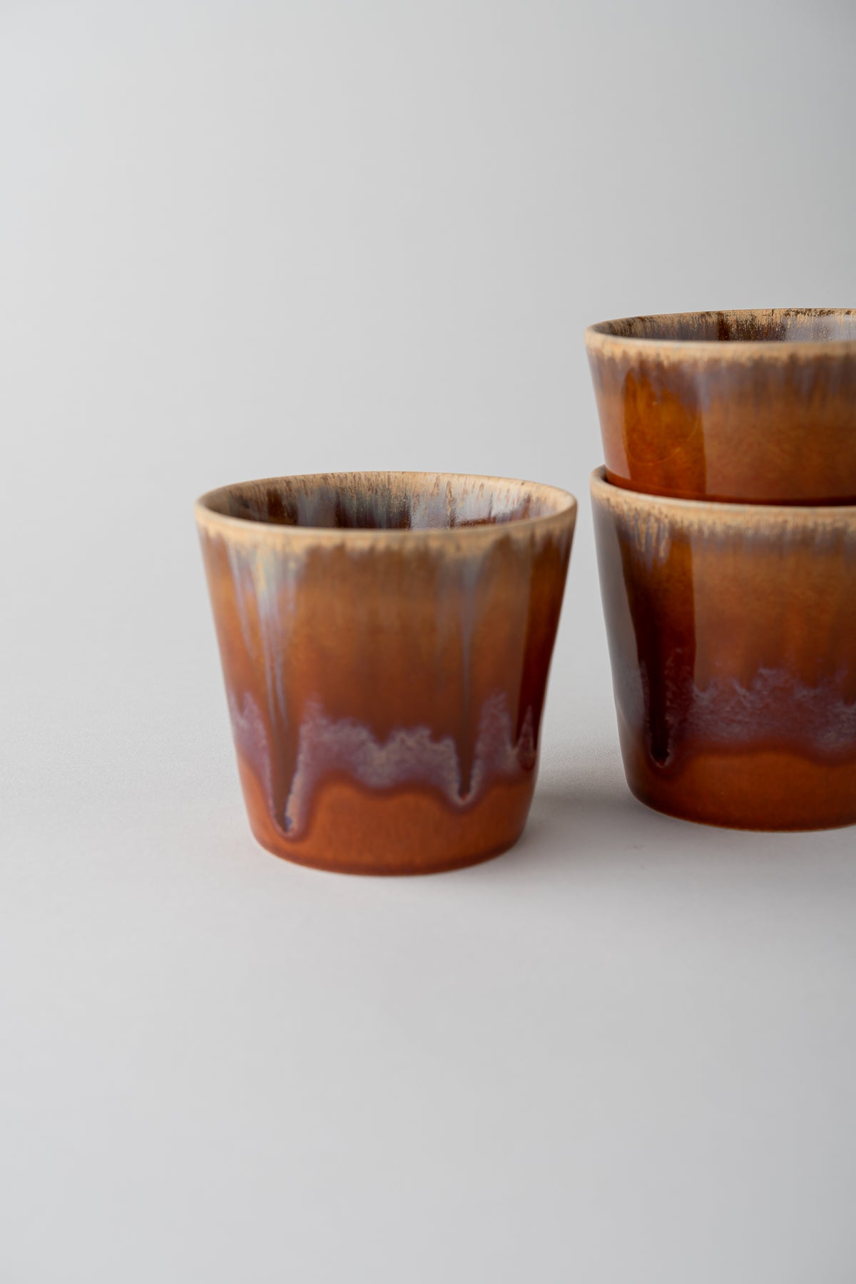 Three small ceramic cups with a glossy brown and amber glaze, featuring a dripping effect near the base, arranged on a light gray background.