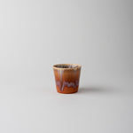 A small ceramic cup with a warm brown and orange body, featuring a dripping glaze pattern in purple and beige, set against a plain light gray background.