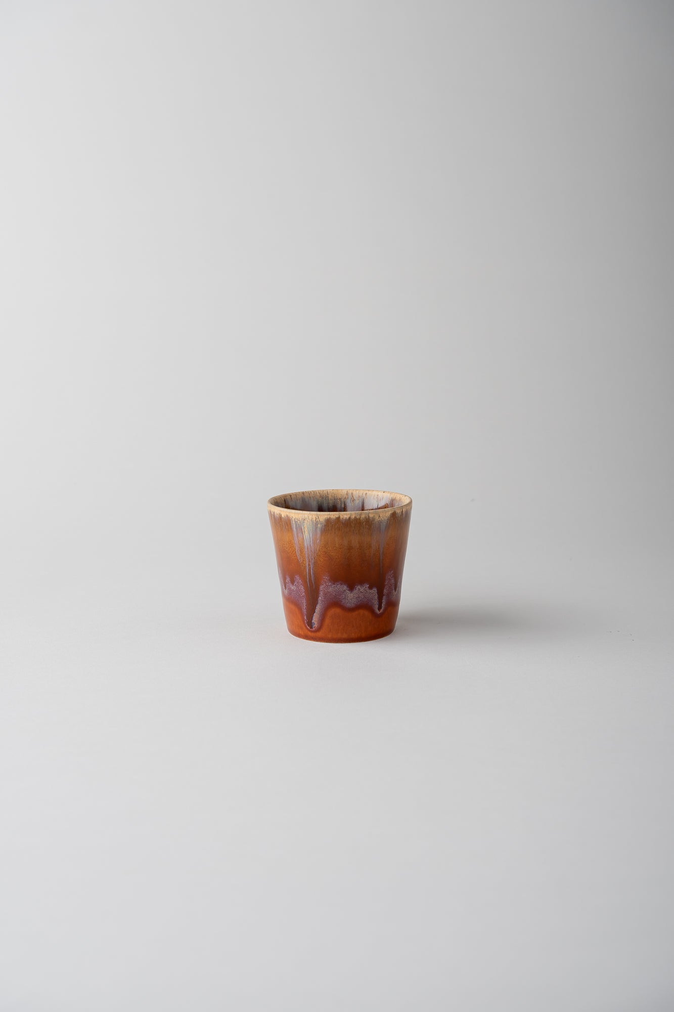 A small ceramic cup with a warm brown and orange body, featuring a dripping glaze pattern in purple and beige, set against a plain light gray background.