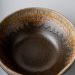 A close-up top-down view of a ceramic bowl with a dark brown interior and a rustic, speckled orange-brown glazed rim, set against a neutral gray background.