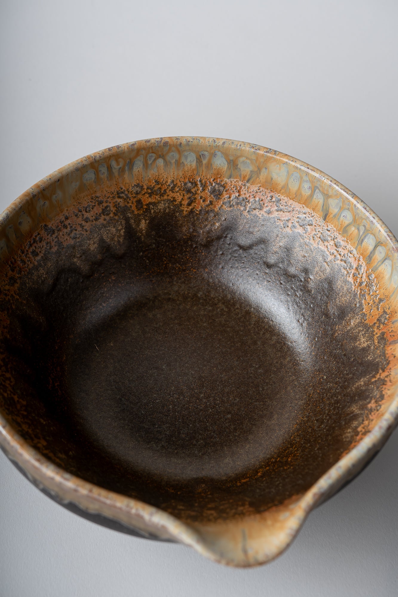 A close-up top-down view of a ceramic bowl with a dark brown interior and a rustic, speckled orange-brown glazed rim, set against a neutral gray background.