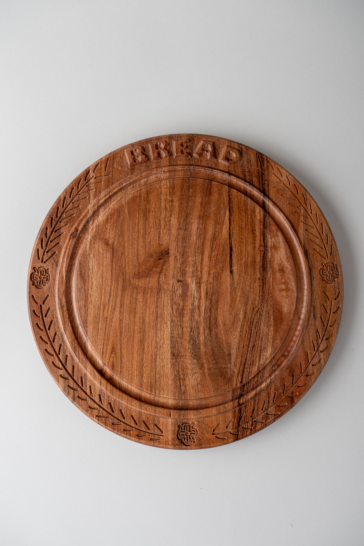 A round wooden cutting board with engraved leaf patterns around the edge and the word 'BREAD' embossed at the top, shown on a light gray background.