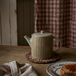 A ribbed ceramic teapot in a light beige glaze sits on a woven coaster on a rustic wooden table, with a plate of jam and a folded cloth napkin nearby against a background of a red-and-white checkered curtain.
