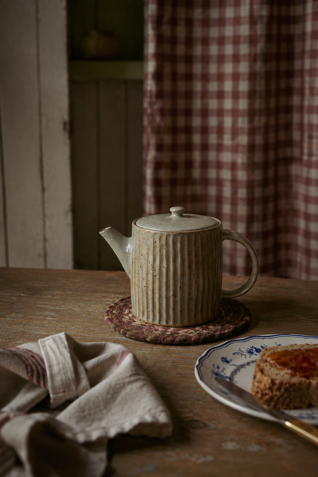 A ribbed ceramic teapot in a light beige glaze sits on a woven coaster on a rustic wooden table, with a plate of jam and a folded cloth napkin nearby against a background of a red-and-white checkered curtain.