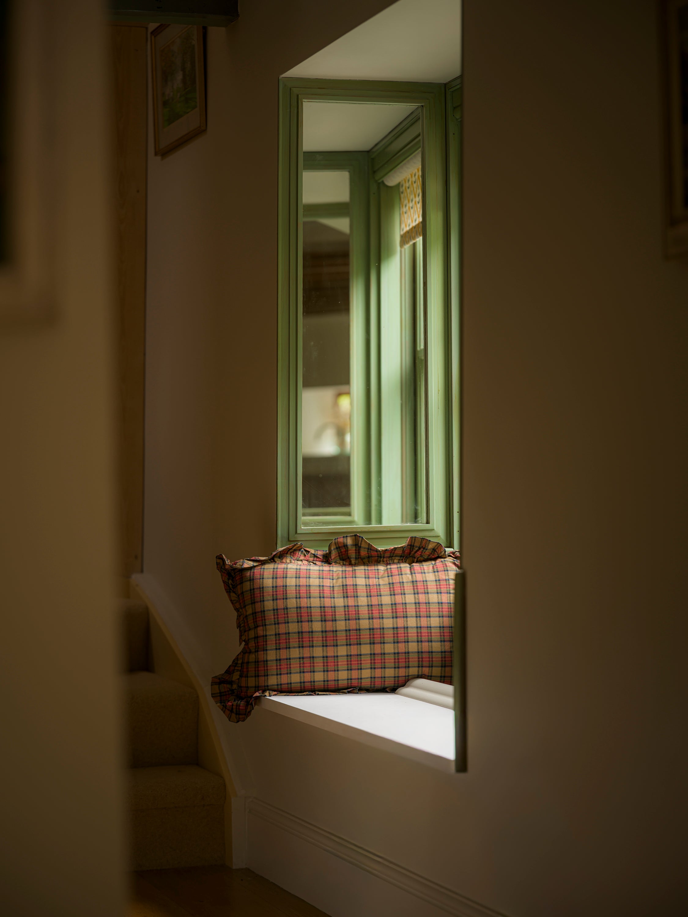 Plaid pillow on a windowsill with a green-framed mirror reflecting a room.