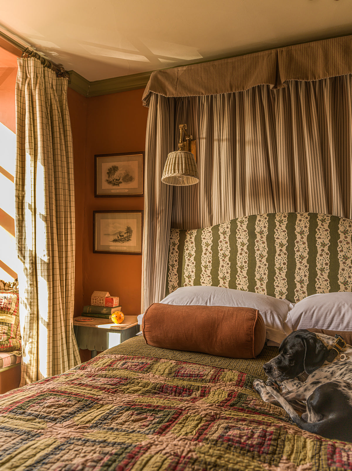 A cozy bedroom scene with a bed dressed in a multicolor quilted coverlet, striped beige and green headboard, striped curtains, warm orange walls, and a black-and-white dog lying on the bed.