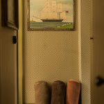 A framed painting of a tall ship hanging on a patterned wallpaper wall in a hallway, with three cylindrical floor cushions in brown and dusty rose standing below.