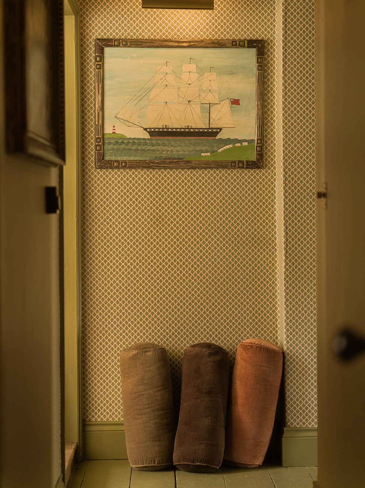 A framed painting of a tall ship hanging on a patterned wallpaper wall in a hallway, with three cylindrical floor cushions in brown and dusty rose standing below.