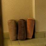 Three cylindrical floor cushions in brown and rust-orange velvet, arranged against a patterned wallpaper wall on a wooden floor.